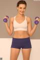 A woman in a white sports bra top and purple shorts holding two purple dumbbells.